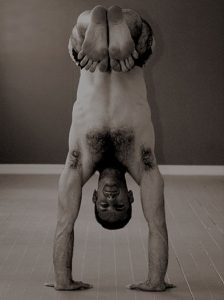 A man performs a handstand with legs bent and feet crossed, balancing upside down on the floor.