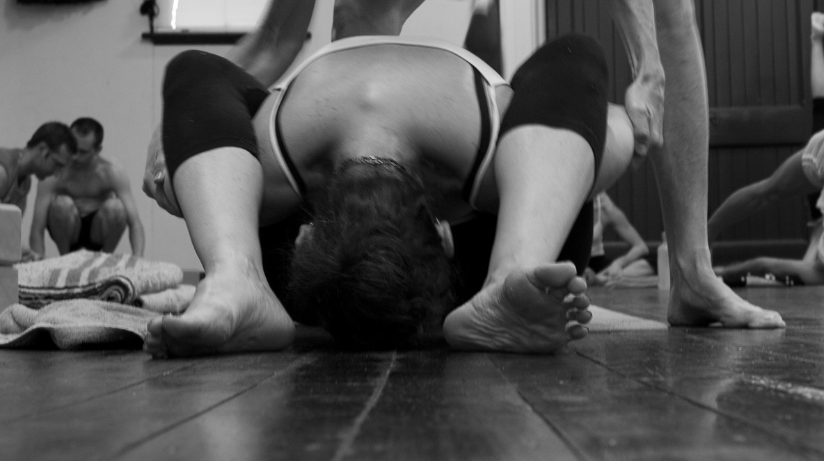 Close-up black and white photo of a yoga practitioner in a deep forward fold, with an instructor assisting the posture.
