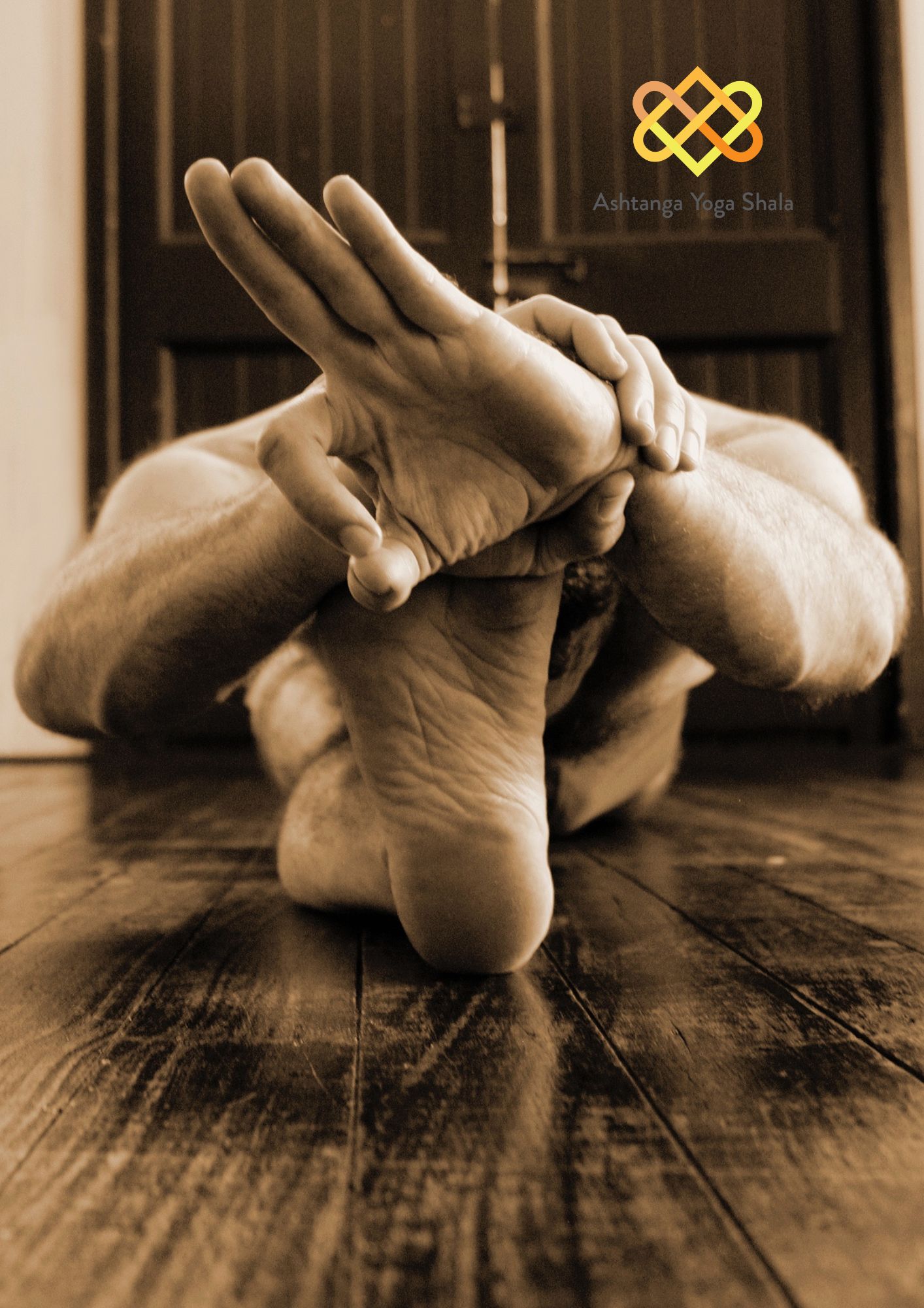 Close-up sepia-toned image of a yoga practitioner in a seated forward bend, reaching toward the feet on a timber floor.
